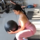 Woman Performing squat exercises with a medicine ball. She’s wearing a pale pink bodysuits and is working in a fitness studio with light bray floors. She has long, black hair that is pulled back in a braid. The view of her is from the side.
