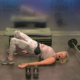 Susan Slater is demonstrating a glute bridge on stage for a fitness livestream. She’s wearing neutral colors leggings and top,and surrounded by fitness equipment, including a step bench, barbell, hand weights and dumbbell weights.